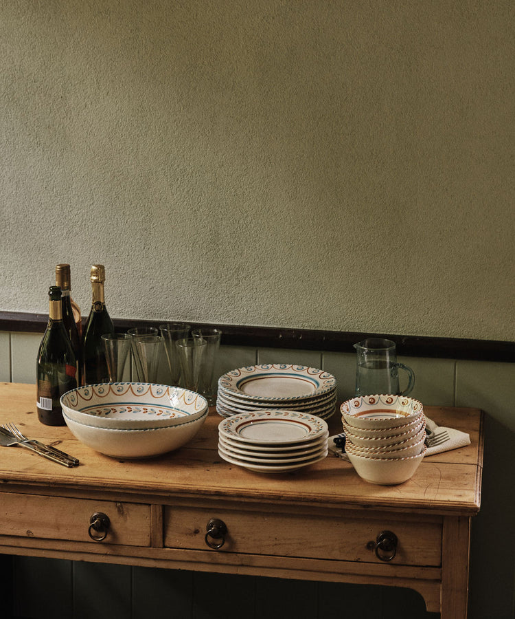 A wooden table holds stacked bowls and plates, including an Ottoline Serving Bowl, drinking glasses, a pitcher, cutlery, and three bottles of sparkling wine against a plain wall.