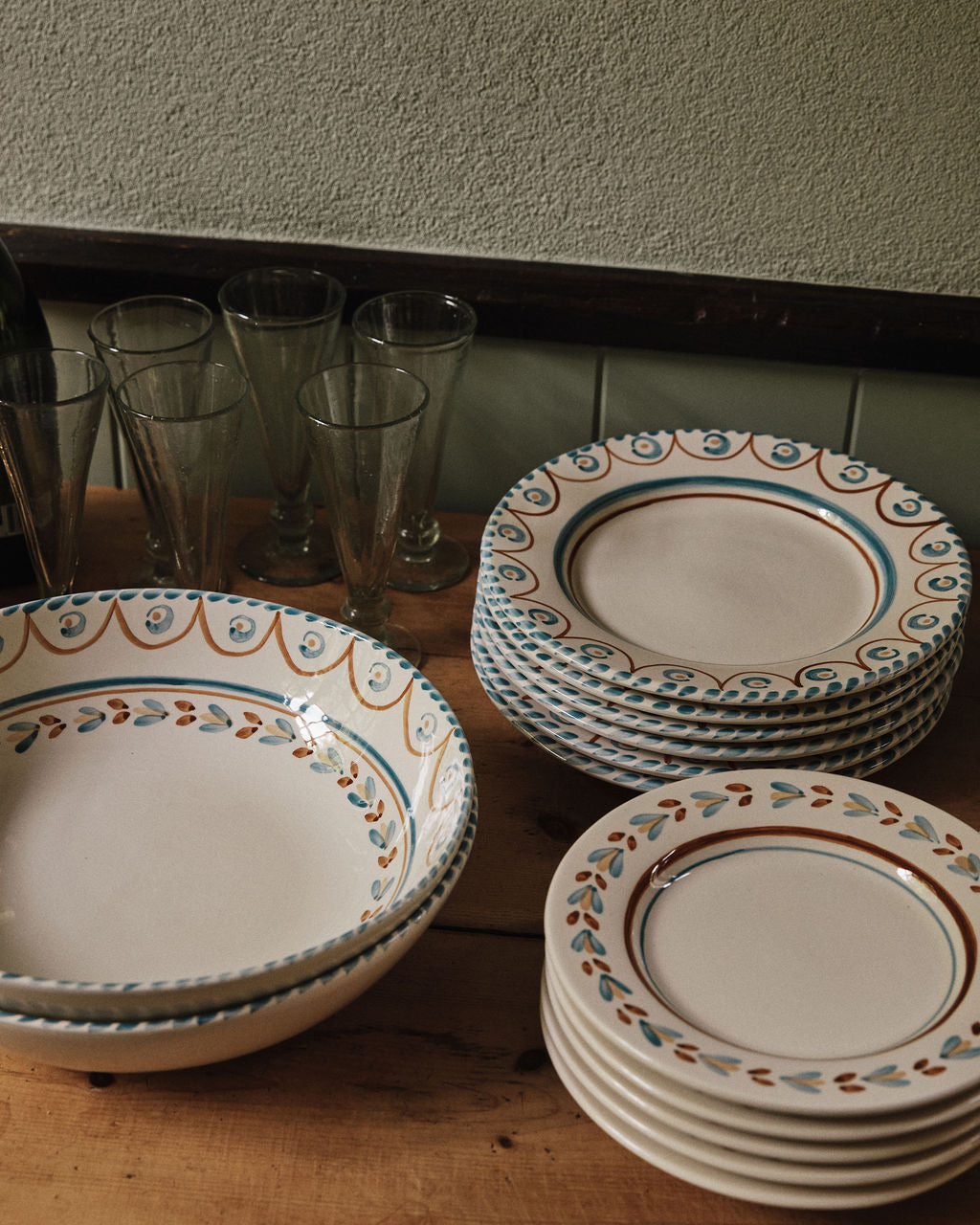 A set of Ottoline Dinner Plates by Tess Newall, hand-painted with blue and orange patterns, is stacked beside four clear drinking glasses on a wooden table.