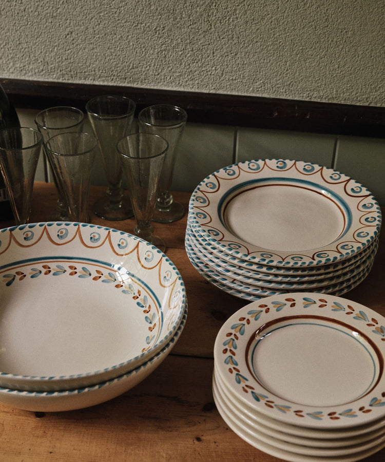 A set of Ottoline Dinner Plates by Tess Newall, hand-painted with blue and orange patterns, is stacked beside four clear drinking glasses on a wooden table.