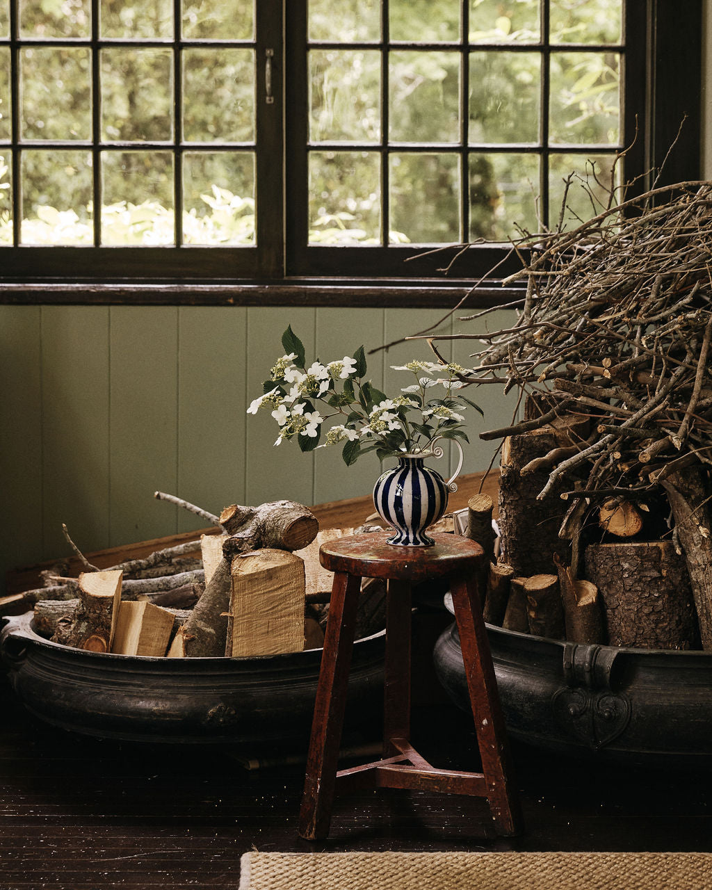 Two large tubs filled with cut firewood and branches sit near a window; a small wooden stool holds the Delphinia Bud Vase filled with white flowers.
