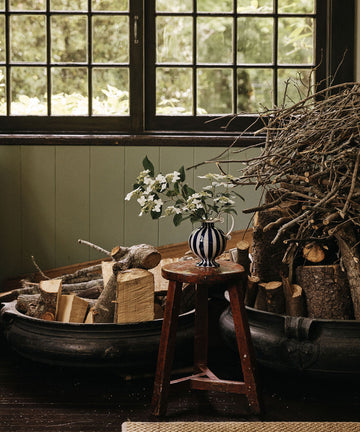 Two large tubs filled with cut firewood and branches sit near a window; a small wooden stool holds the Delphinia Bud Vase filled with white flowers.