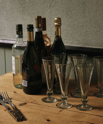 A set of six Elodie Champagne Glasses is arranged on a wooden table with several empty wine and champagne bottles, two forks, and two knives.
