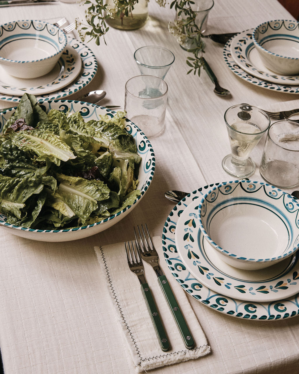 A table set with patterned plates, bowls, glasses, and utensils rests on a cream tablecloth, accented by Filial Napkin, Set of 6 — each textured napkin folded neatly beside every setting. In the center sits a large bowl of leafy salad and two glasses of white wine, creating an inviting scene for a shared meal.