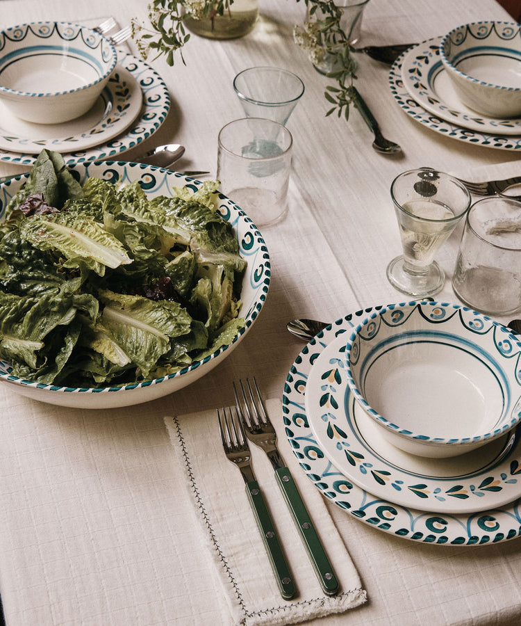 A table set with patterned plates, bowls, glasses, and utensils rests on a cream tablecloth, accented by Filial Napkin, Set of 6 — each textured napkin folded neatly beside every setting. In the center sits a large bowl of leafy salad and two glasses of white wine, creating an inviting scene for a shared meal.