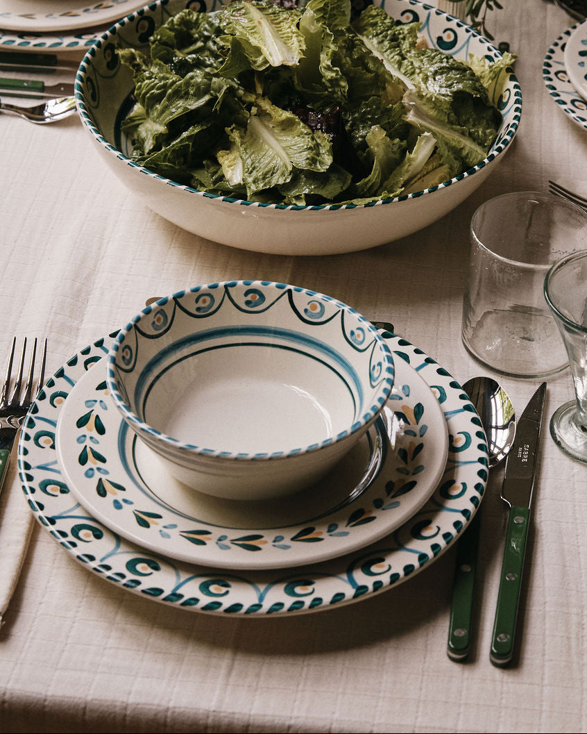 A dining table set with patterned plates, Ottoline Cereal Bowls (set of 4), silverware, clear glasses, and a large salad bowl filled with leafy greens on a light tablecloth.