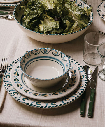 A dining table set with patterned plates, Ottoline Cereal Bowls (set of 4), silverware, clear glasses, and a large salad bowl filled with leafy greens on a light tablecloth.