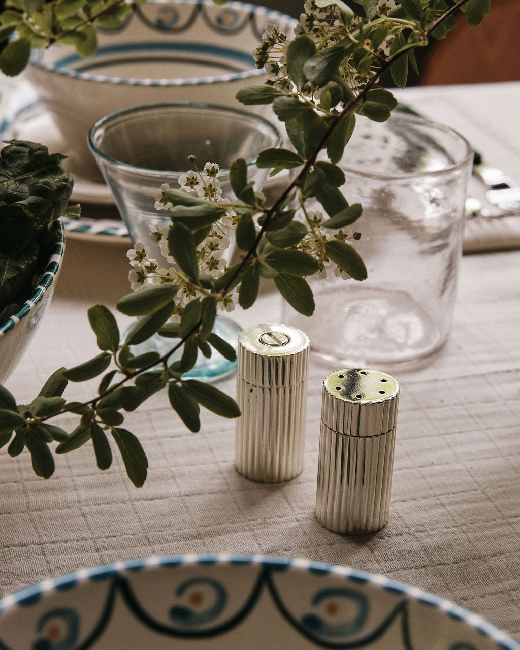 The Sophia Pepper Mill and Salt Shaker, a testament to exquisite Italian craftsmanship, sit elegantly on a set dining table adorned with glassware, patterned dishes, and green leafy branches as decoration.