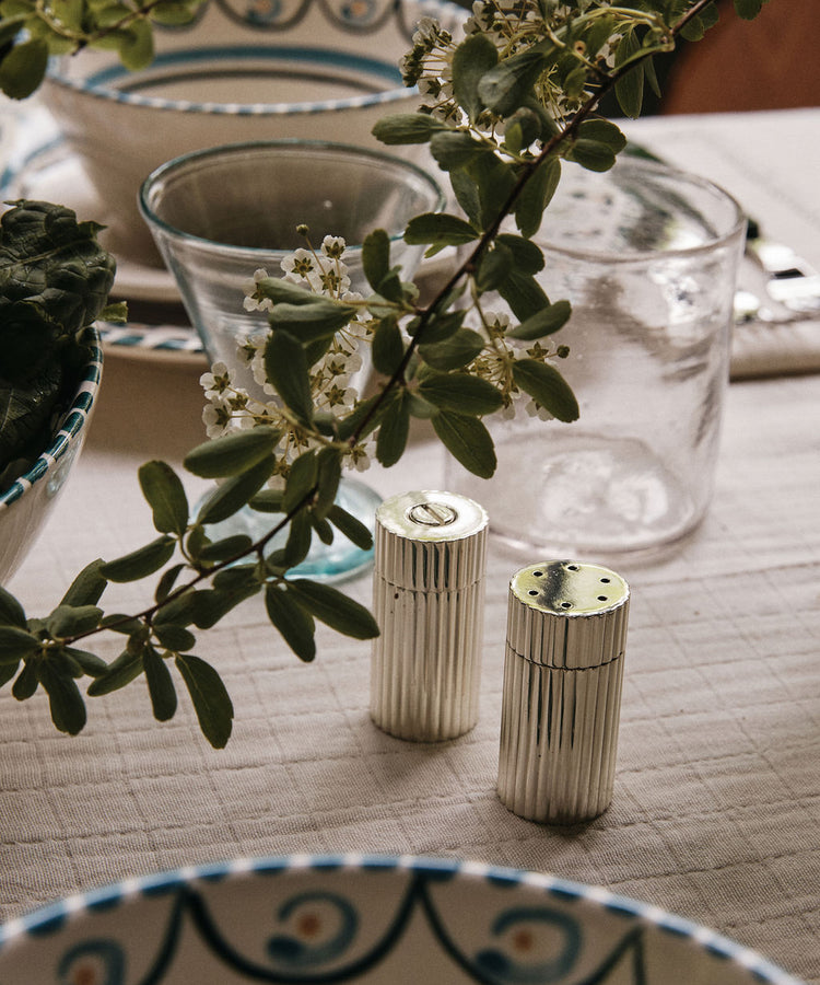 The Sophia Pepper Mill and Salt Shaker, a testament to exquisite Italian craftsmanship, sit elegantly on a set dining table adorned with glassware, patterned dishes, and green leafy branches as decoration.