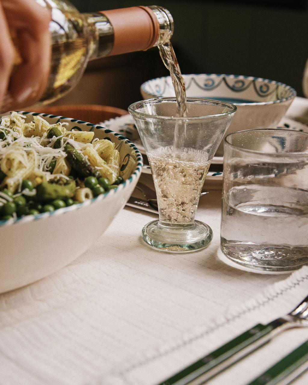 A hand pours white wine into an Elodie Footed Wine Glass next to a bowl of pasta with vegetables and cheese on a set dining table.