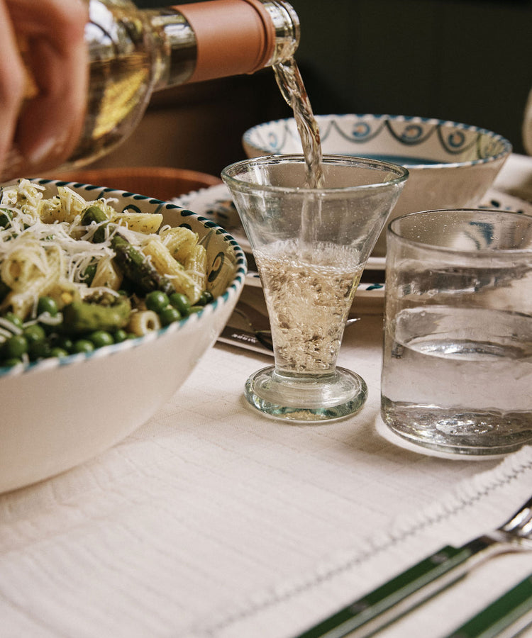 A hand pours white wine into an Elodie Footed Wine Glass next to a bowl of pasta with vegetables and cheese on a set dining table.