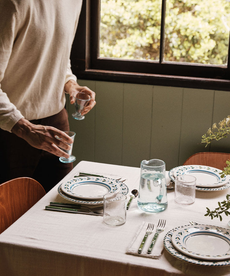 A person sets drinking glasses, handblown in Damascus, on a table with a white tablecloth, plates, cutlery, and the Elodie Pitcher filled with water near a window with greenery outside.