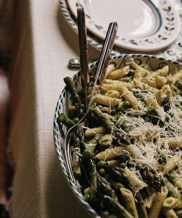 A bowl of penne pasta with asparagus, peas, and grated cheese sits on the table in an Ottoline Serving Bowl, next to plates and cutlery.