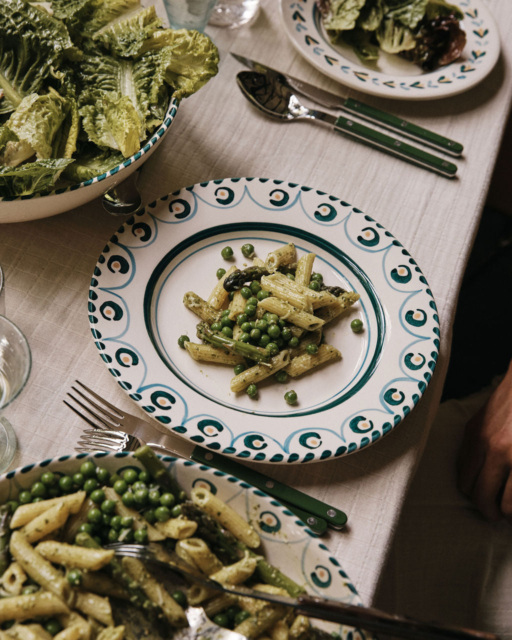 A plate of penne pasta with green peas is served on an Ottoline Dinner Plate (set of 4), surrounded by salad and utensils on a beige tablecloth.