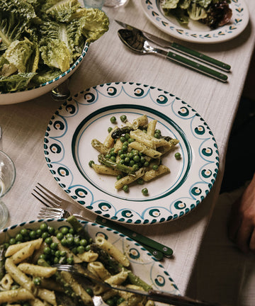 A plate of penne pasta with green peas is served on an Ottoline Dinner Plate (set of 4), surrounded by salad and utensils on a beige tablecloth.
