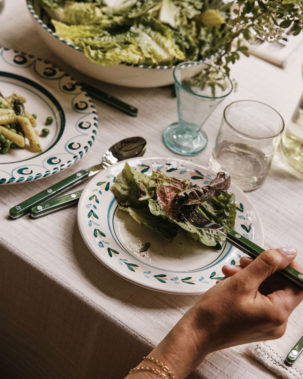 A person holds a fork with mixed greens over an Ottoline Small Plate (Set of 4) on a set dining table, complete with a bowl of lettuce, pasta, water, and glassware.