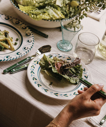 A person holds a fork with mixed greens over an Ottoline Small Plate (Set of 4) on a set dining table, complete with a bowl of lettuce, pasta, water, and glassware.