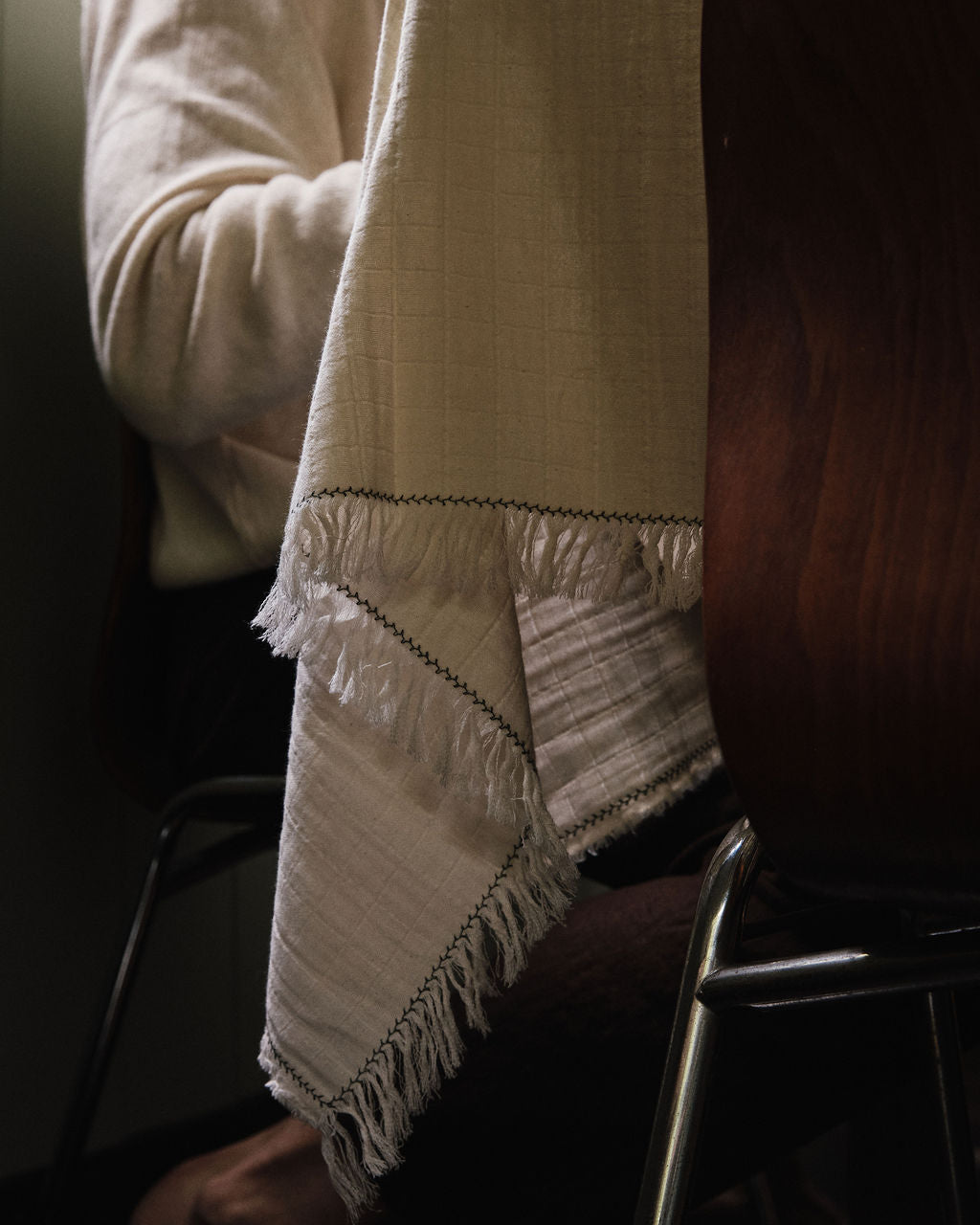 A person sits at a table covered with the Filial Tablecloth, which is white with fringes and features a delicate green branch stitch; only their upper body and part of the chair can be seen.