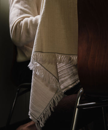 A person sits at a table covered with the Filial Tablecloth, which is white with fringes and features a delicate green branch stitch; only their upper body and part of the chair can be seen.