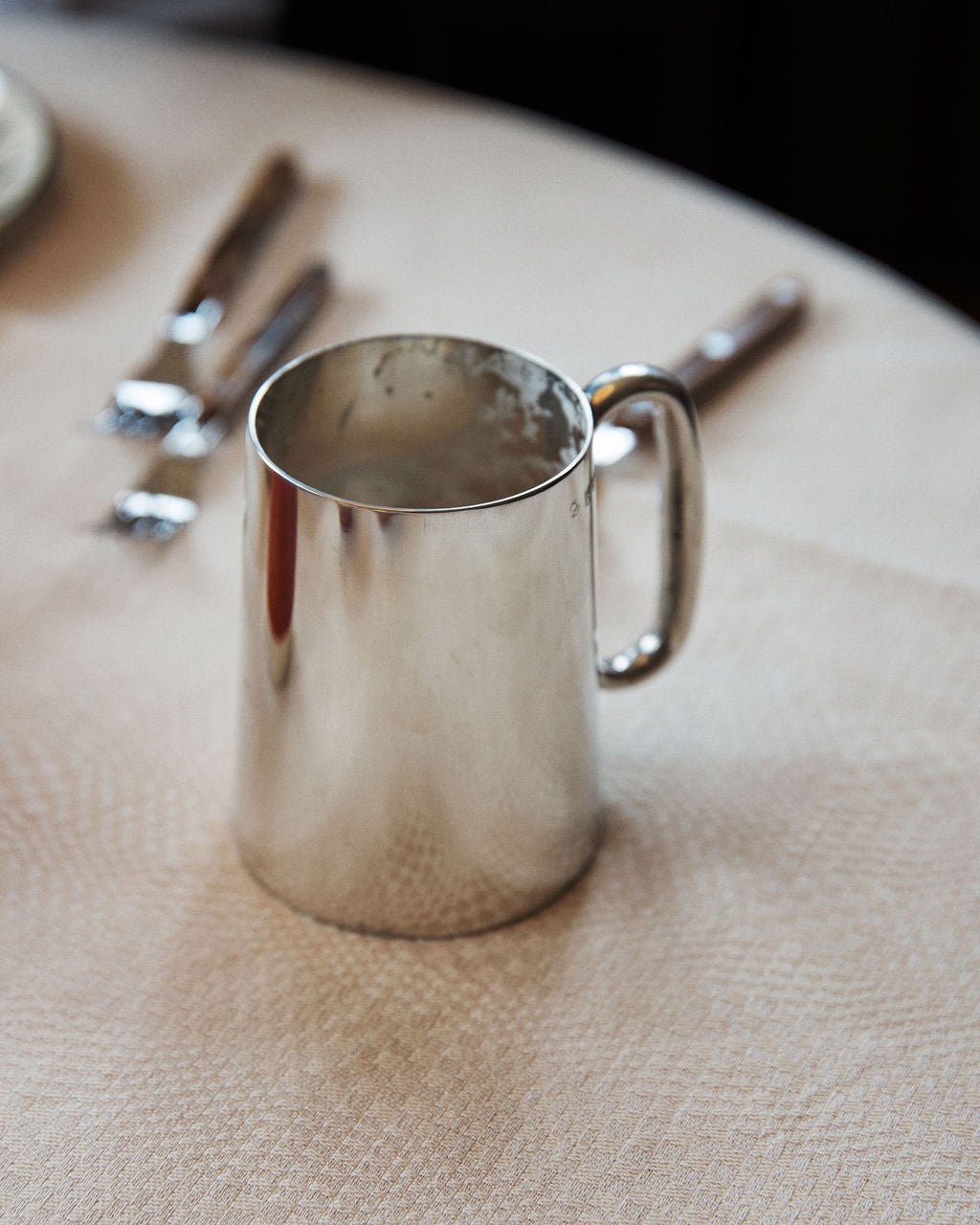 The Harry Tankard, a shiny tarnish-free metal mug inspired by pewter beer tankards, rests on a light-colored tablecloth with cutlery visible in the background.