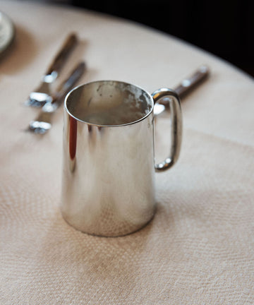 The Harry Tankard, a shiny tarnish-free metal mug inspired by pewter beer tankards, rests on a light-colored tablecloth with cutlery visible in the background.