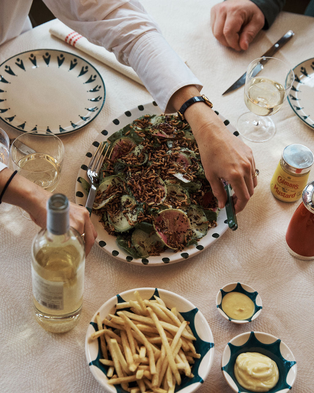 A table set for a meal features hand-glazed ceramic plates, glasses, condiments, wine, and the Nina Oval Serving Platter holding sliced meat and greens; two people are seated.