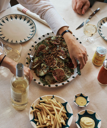 A table set for a meal features hand-glazed ceramic plates, glasses, condiments, wine, and the Nina Oval Serving Platter holding sliced meat and greens; two people are seated.