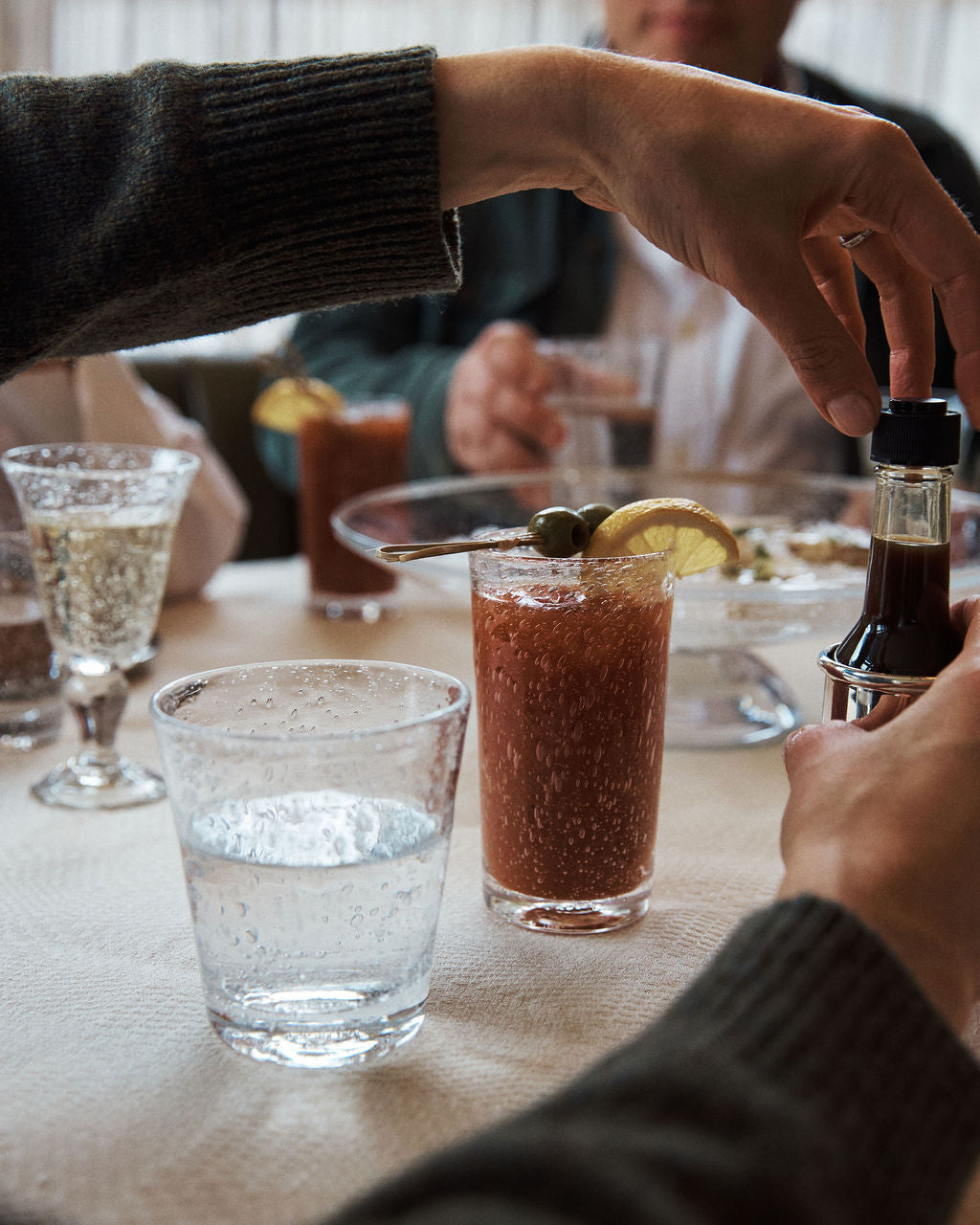 A person prepares a Bloody Mary cocktail in a Lauren Juice Glass (Set of 2) at a restaurant table with sparkling water, wine, and food, while others sit nearby.