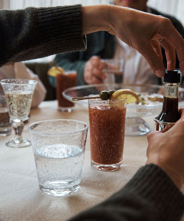 A person prepares a Bloody Mary cocktail in a Lauren Juice Glass (Set of 2) at a restaurant table with sparkling water, wine, and food, while others sit nearby.
