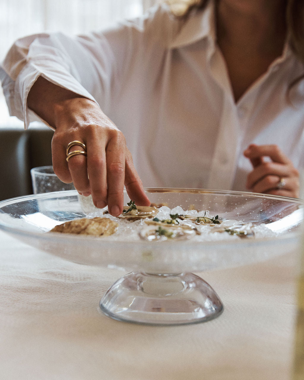 A person in a white shirt reaches for an oyster on the Bubble Glass Grape Tray, which is filled with ice and herbs, adding elegance to the table.