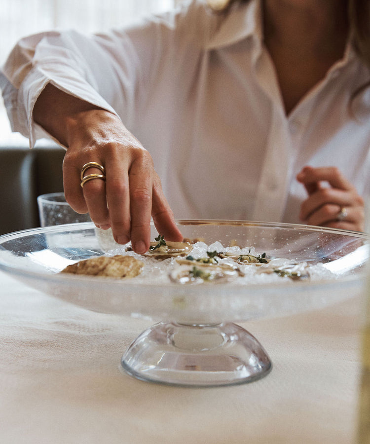 A person in a white shirt reaches for an oyster on the Bubble Glass Grape Tray, which is filled with ice and herbs, adding elegance to the table.