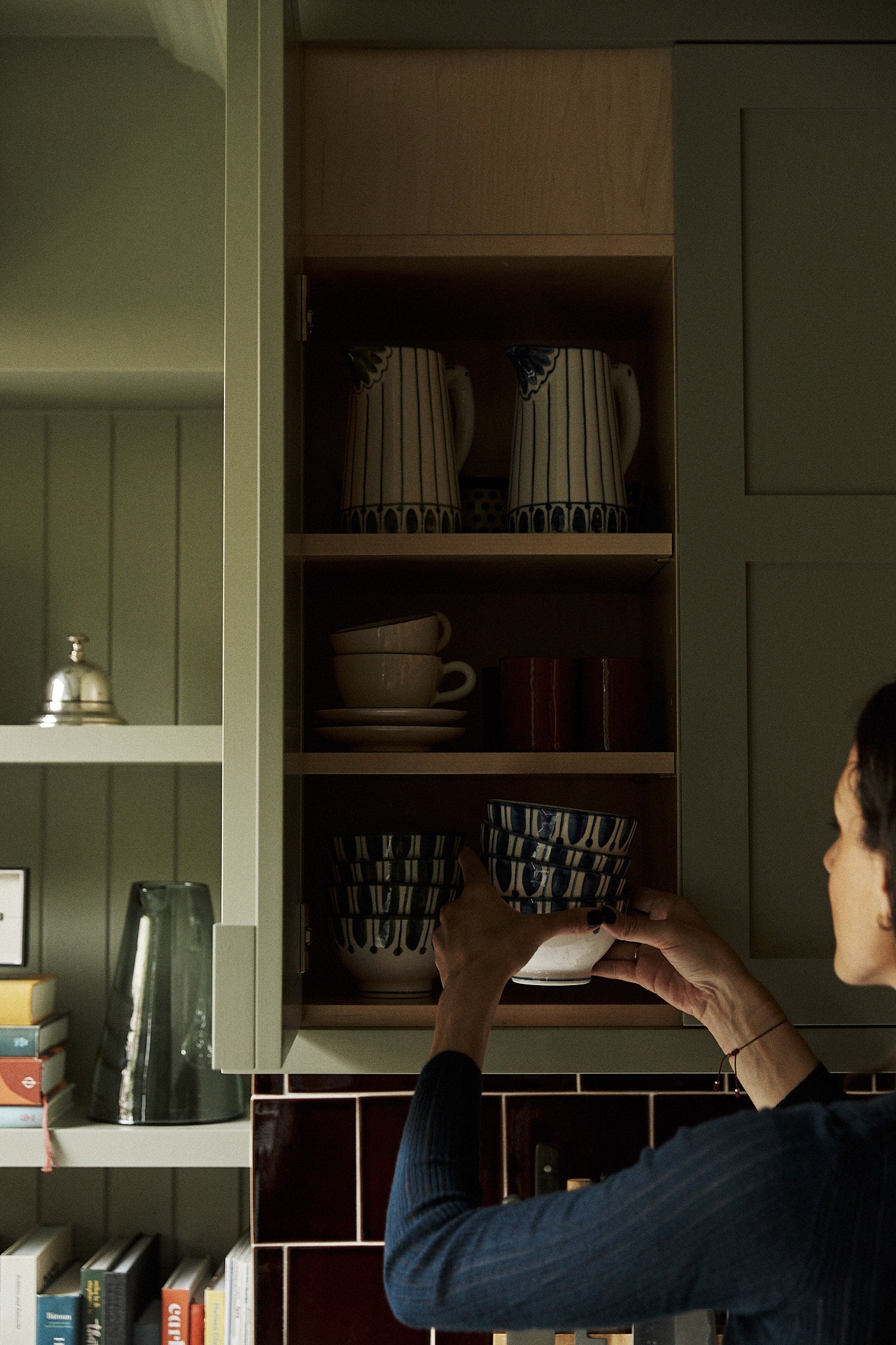 A person places a Jorge Cereal Bowl, Set of 4, on a kitchen shelf inside a cabinet filled with elegant tableware and matching pitchers.