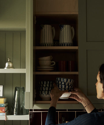 A person places a Jorge Cereal Bowl, Set of 4, on a kitchen shelf inside a cabinet filled with elegant tableware and matching pitchers.