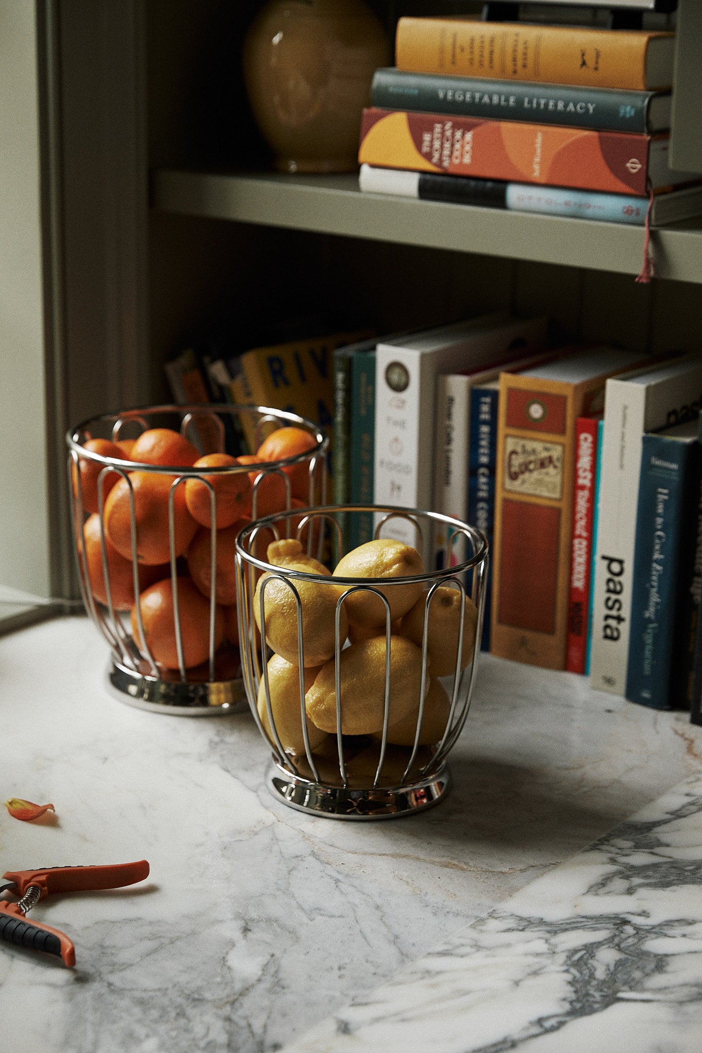 Two Spicchio Citrus Baskets filled with oranges and pears sit on a marble countertop, adding sculptural flair, while books and a vase are displayed on the shelves in the background.