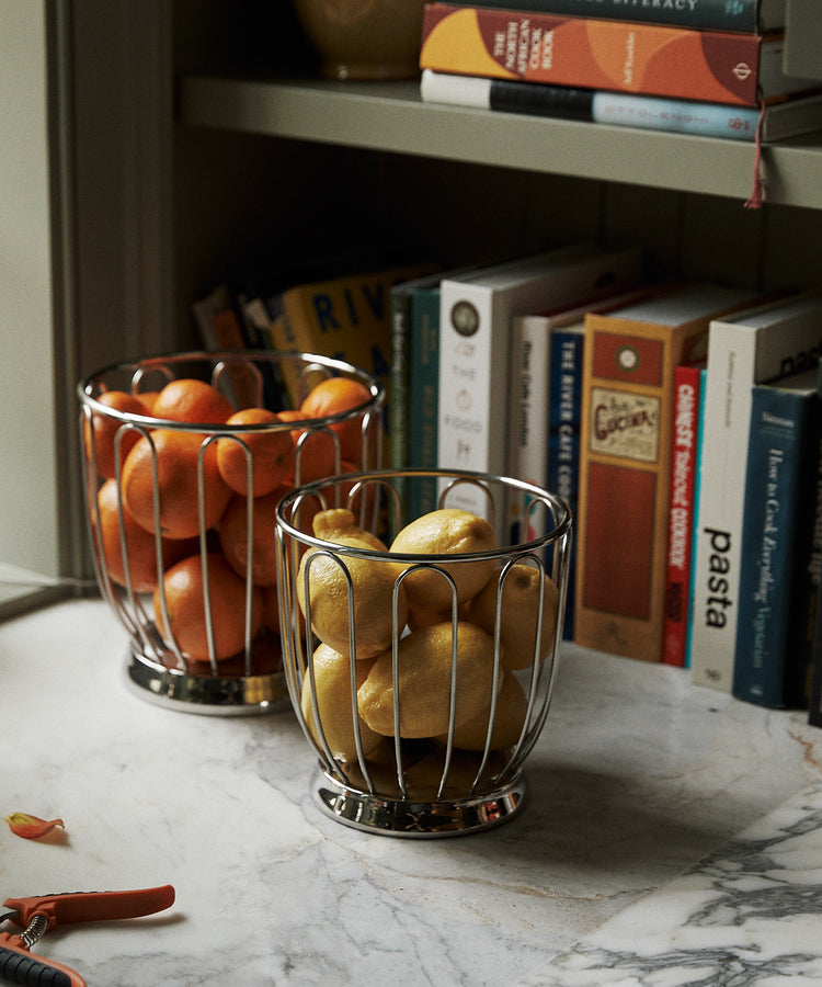 Two Spicchio Citrus Baskets filled with oranges and pears sit on a marble countertop, adding sculptural flair, while books and a vase are displayed on the shelves in the background.