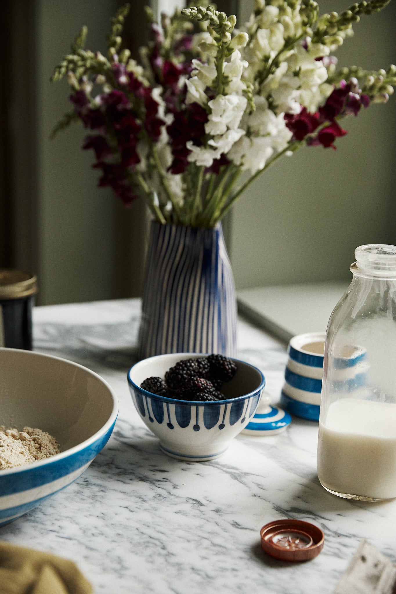 A marble countertop with a Jorge Cereal Bowl, Set of 4—one filled with flour, alongside blackberries, a bottle of milk, a jar lid, and a vase of flowers in the background.