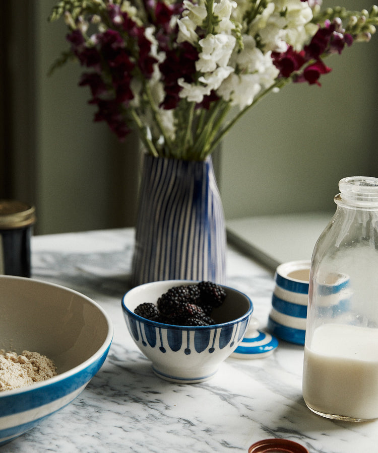 A marble countertop with a Jorge Cereal Bowl, Set of 4—one filled with flour, alongside blackberries, a bottle of milk, a jar lid, and a vase of flowers in the background.