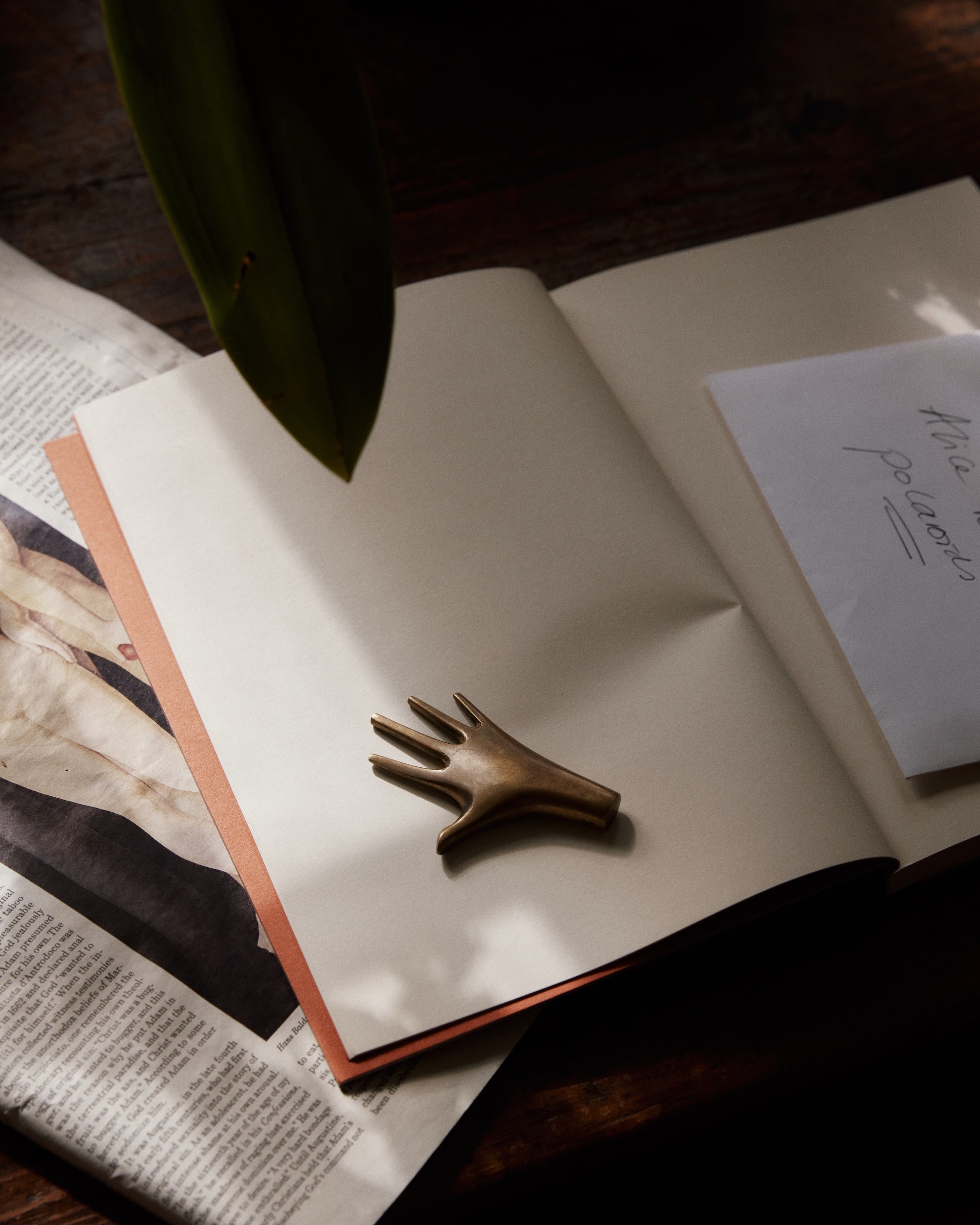The Hand Paperweight, crafted from solid brass by the Auböck workshop, rests on an open notebook beside folded paper in gentle natural light.