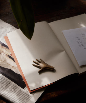 The Hand Paperweight, crafted from solid brass by the Auböck workshop, rests on an open notebook beside folded paper in gentle natural light.