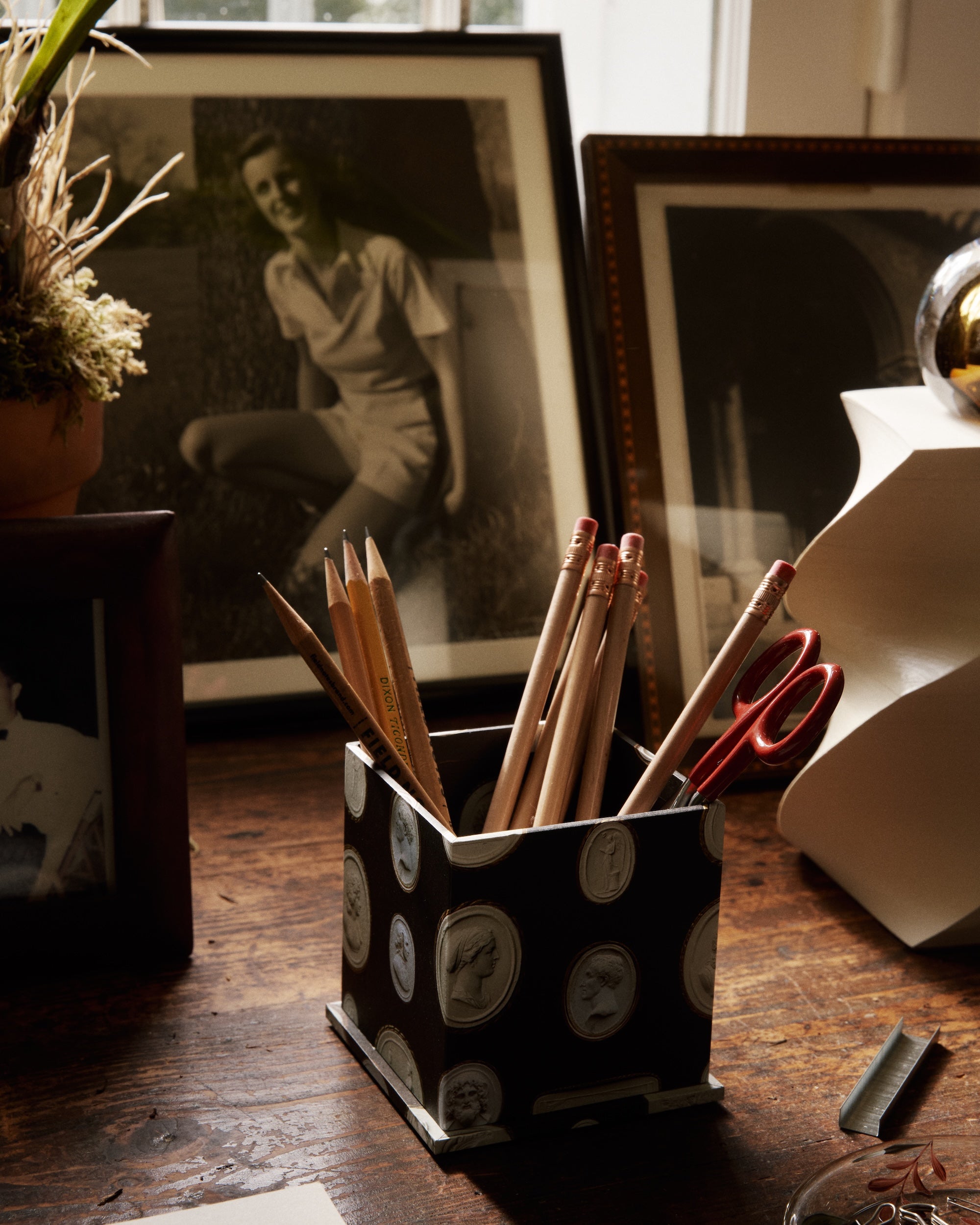 The Intaglio Pencil Pot filled with pencils and scissors sits on a wooden desk surrounded by book boards, photos, papers, a lamp, and a round decorative object.