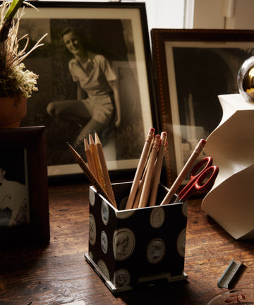 The Intaglio Pencil Pot filled with pencils and scissors sits on a wooden desk surrounded by book boards, photos, papers, a lamp, and a round decorative object.