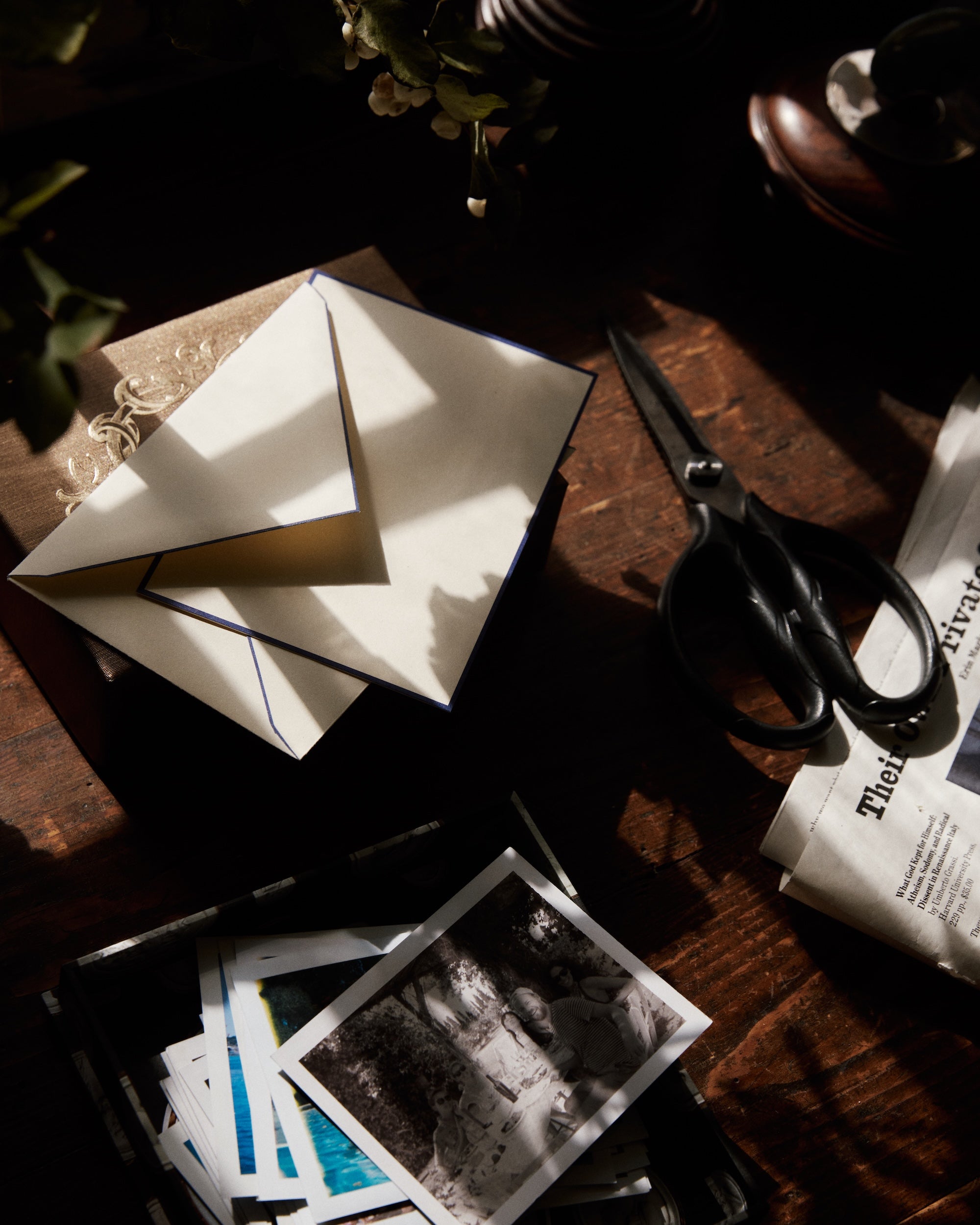 A Bordered Correspondence Card & Envelope, Set of 25 rests on a book near scissors, photos, and a newspaper on a sunlit wooden table.