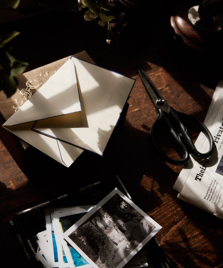 A Bordered Correspondence Card & Envelope, Set of 25 rests on a book near scissors, photos, and a newspaper on a sunlit wooden table.