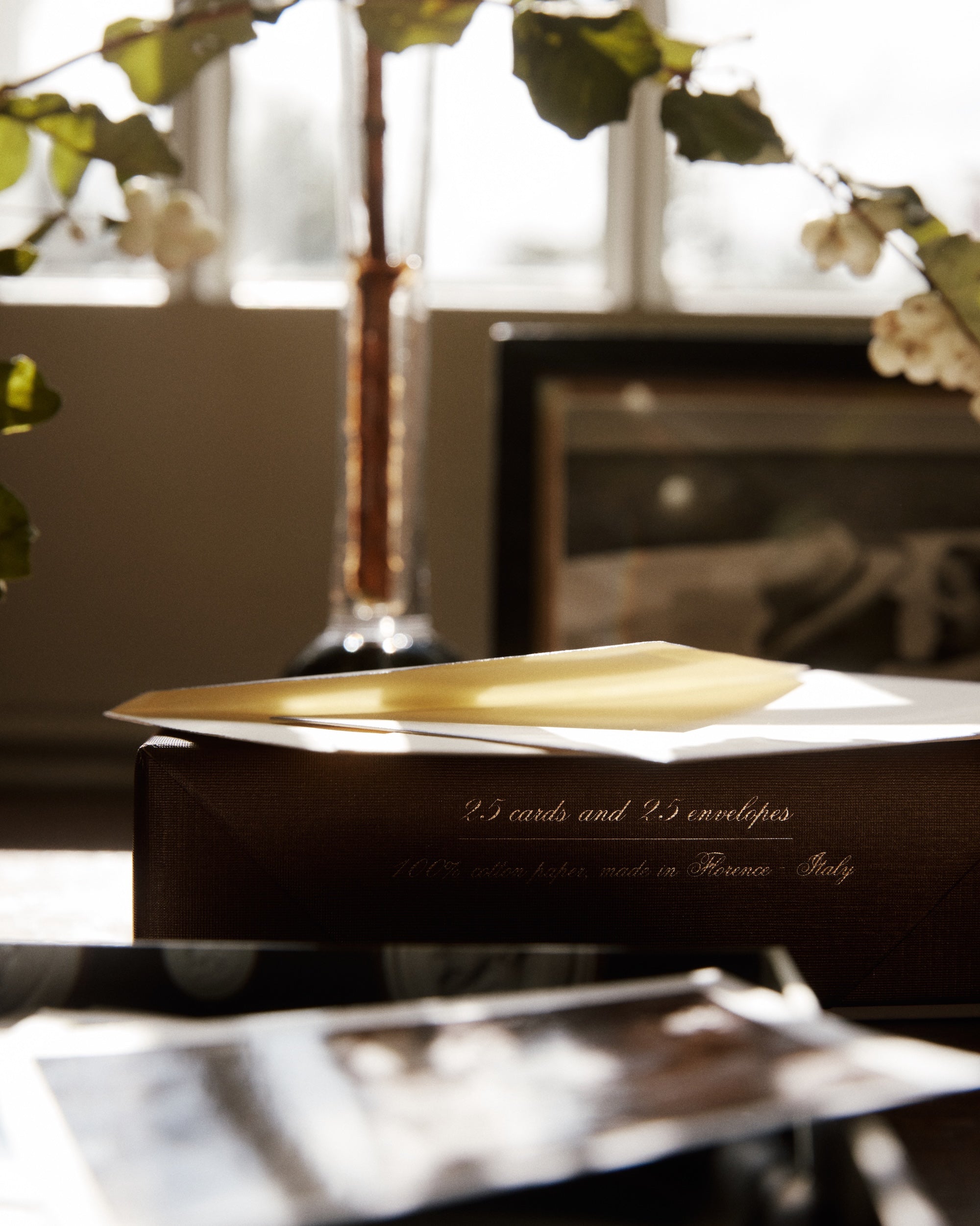 A yellow envelope sits on a box labeled Bordered Correspondence Card & Envelope, Set of 25, next to blurred photos and a vase with branches in soft daylight.
