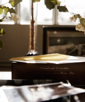 A yellow envelope sits on a box labeled Bordered Correspondence Card & Envelope, Set of 25, next to blurred photos and a vase with branches in soft daylight.