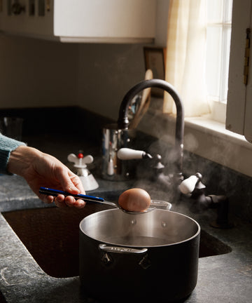 A hand lifts a brown egg with a spoon from a steaming pot, using the Bistrot 5 Piece Cutlery Set with navy blue handles by the kitchen sink and window.