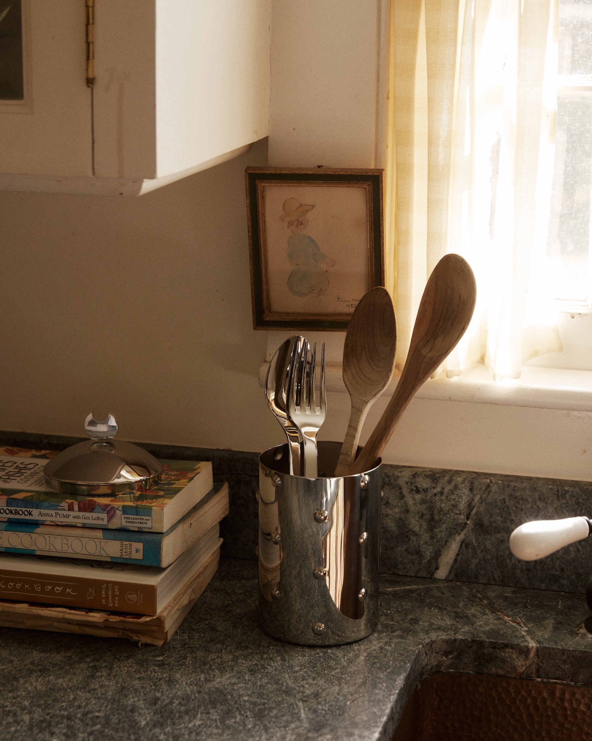 The Kalisto Kitchen Box offers stylish stainless steel storage for utensils on your counter beside cookbooks, a window, and framed art.