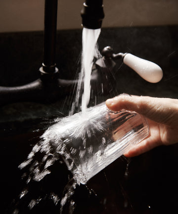 A hand rinses a Teo Ridged Water Glass, Set of 4 under a kitchen faucet, with water splashing over the sides.