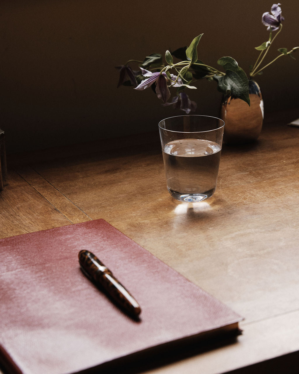 A closed red notebook with a pen rests on a wooden desk next to an Arc Tumbler, Set of 4 glass of water and a small vase holding purple flowers.
