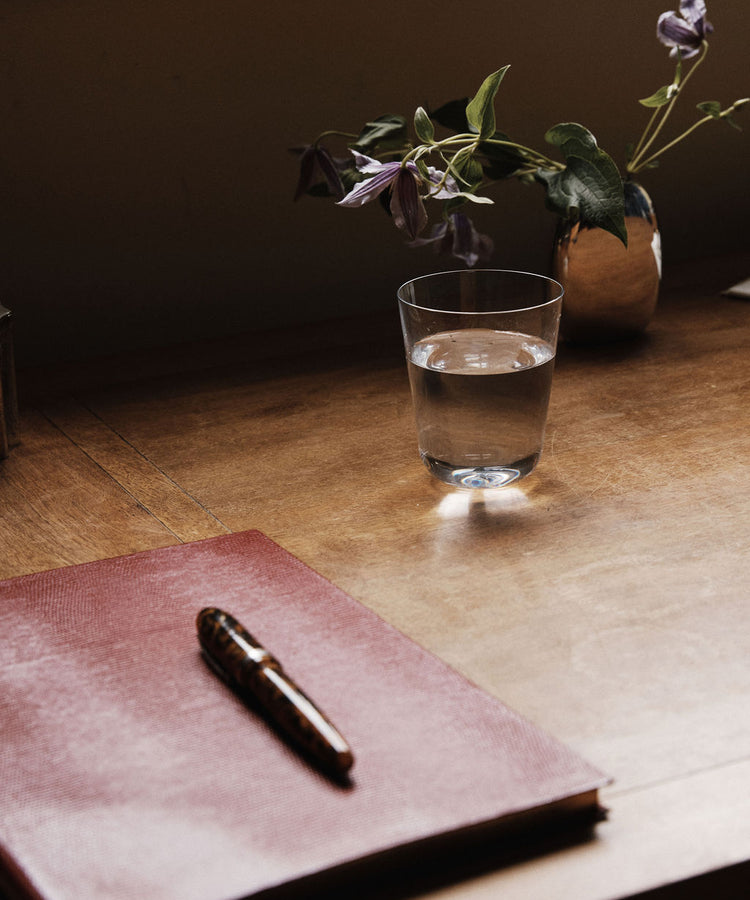 A closed red notebook with a pen rests on a wooden desk next to an Arc Tumbler, Set of 4 glass of water and a small vase holding purple flowers.