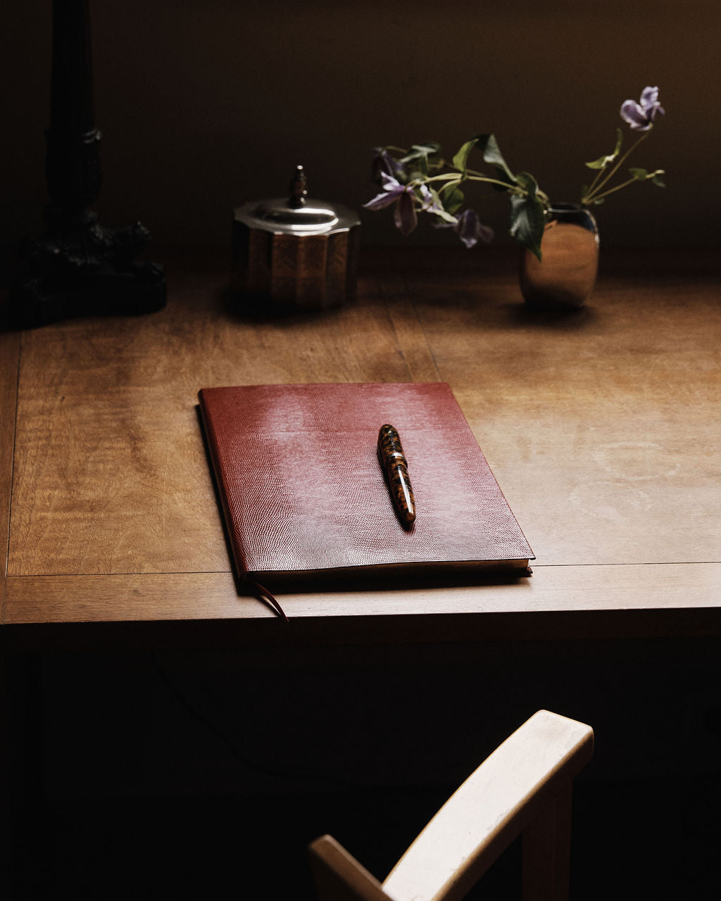 The Red Notebook with a lizard-embossed cover and a pen rests on a wooden desk near a small vase of purple flowers and a decorative container.